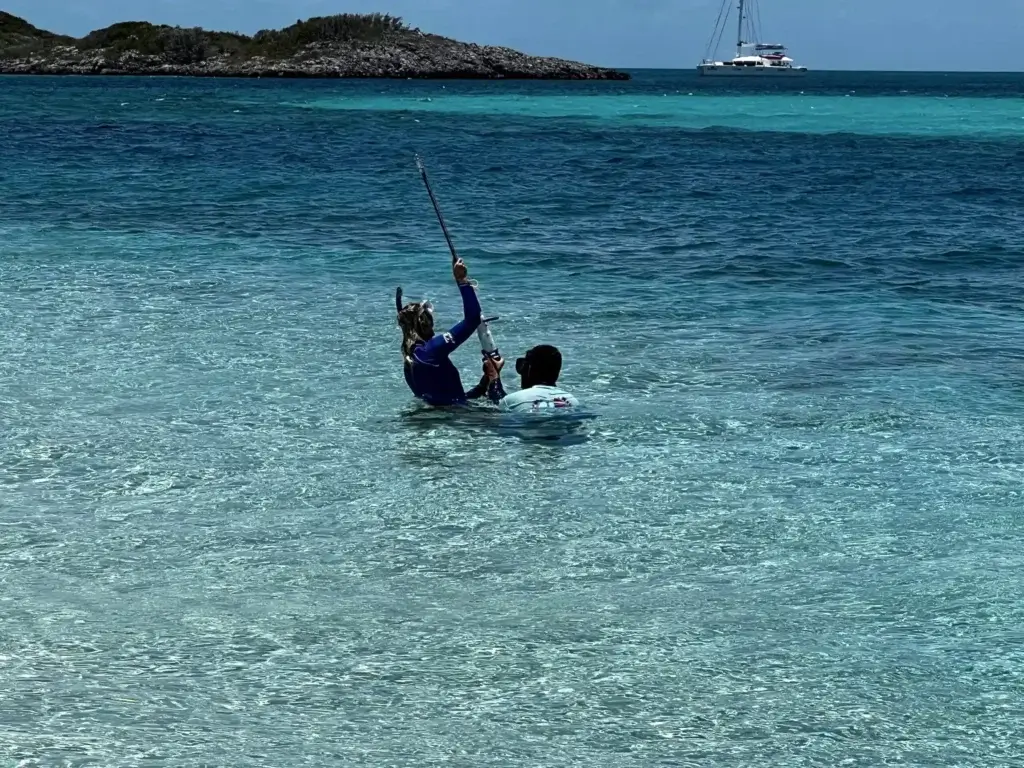 Two individuals fishing in clear, shallow water using traditional spear fishing, prompting consideration of its sustainability compared to modern methods and overfishing.